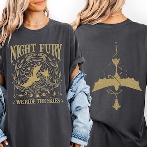 May include: Dark gray t-shirt with a gold graphic featuring a dragon and the text "NIGHT FURY ISLE OF BERK WE RIDE THE SKIES." The back of the shirt has a gold dragon design. The shirt is a comfortable, casual style.