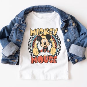 May include: White t-shirt featuring a retro Mickey Mouse graphic with the text "Mickey Mouse". The shirt is layered under a blue denim jacket. The graphic includes a circular design with stars and a checkered pattern.