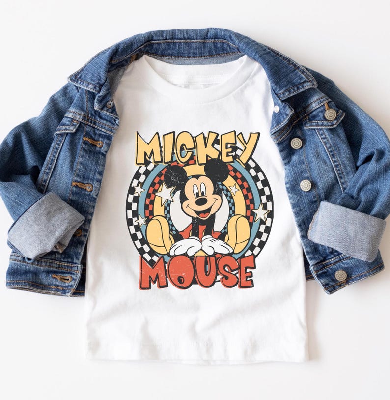 May include: White t-shirt featuring a retro Mickey Mouse graphic with the text "Mickey Mouse". The shirt is layered under a blue denim jacket. The graphic includes a circular design with stars and a checkered pattern.
