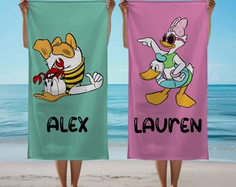 May include: Two beach towels, one teal and one pink, are held up against a beach backdrop. The teal towel features Donald Duck with a crab, and the name "ALEX". The pink towel features Daisy Duck and Donald Duck, with the name "LAUREN".