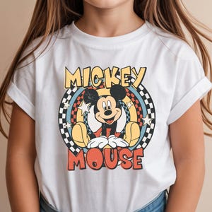 May include: White t-shirt featuring a retro Mickey Mouse graphic. The design includes the words "Mickey Mouse" in a vintage font, with Mickey in the center. The graphic has a circular design with stars and a checkered pattern.