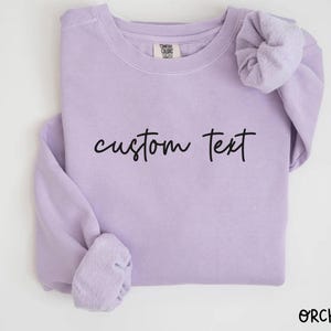 May include: A light purple sweatshirt with the words "custom text" in black script. The sweatshirt has long sleeves and a crew neck. The word "ORCHID" is printed in the bottom right corner.