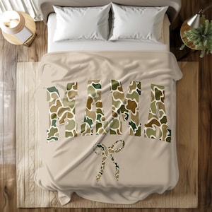 May include: A beige blanket featuring the word "MAMA" in large letters with a camouflage design. A camouflage bow is below the word. The blanket is on a bed with white pillows and a wooden frame.