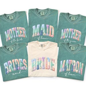 May include: A collection of six t-shirts in various colors, including green and cream. Each shirt features a unique text design, such as "Mother of the Bride," "Maid of Honor," and "Bride." The text is in a patchwork style, with a delicate necklace.