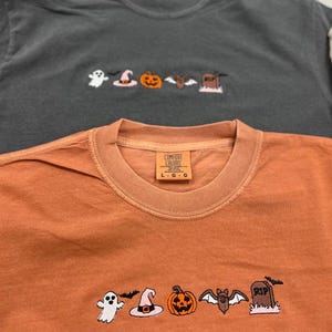 Puede incluir: Dos camisetas de manga larga, una gris antracita y otra naranja, con diseños bordados con temática de Halloween. Los diseños incluyen un fantasma, un sombrero de bruja, una calabaza, un murciélago y una lápida. La camiseta naranja tiene una etiqueta Comfort Colors.