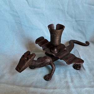 Puede incluir: Un candelabro con forma de dragón de color bronce. El dragón tiene las alas extendidas, una larga cola y un portavelas en la espalda. El candelabro es de metal y está sobre una superficie azul claro.