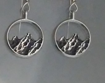 Puede incluir: Un par de pendientes de plata con un diseño de cordillera. Las montañas están silueteada sobre un fondo blanco.