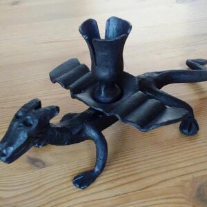 May include: Black metal dragon-shaped candle holder. The candle holder features a dragon with outstretched wings and a cup-shaped holder for a single candle. The dragon is detailed with scales and claws. The candle holder is approximately 6 inches long.