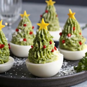 Peut inclure: Des œufs farcis en forme de sapins de Noël. La garniture verte est surmontée d'une étoile jaune et de morceaux de poivron rouge. Les œufs sont disposés sur une assiette sombre, une entrée festive pour les fêtes.