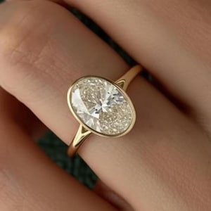 Könnte beinhalten: Ein goldener Ring mit einem oval geschliffenen Diamanten. Der Diamant ist in einer Zargenfassung gefasst, und das Band ist aus schlichtem, poliertem Gold. Der Ring wird auf einer Hand vor grünem Hintergrund gezeigt.