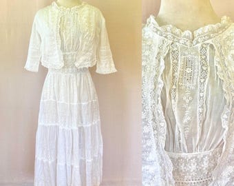 May include: A white vintage dress and bolero jacket set. The dress features a tiered skirt and intricate lace detailing. The bolero jacket has a high collar and short sleeves, also with lace embellishments. The ensemble is displayed on a mannequin.