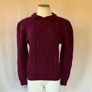 Puede incluir: Un suéter burdeos con cuello y mangas largas. El suéter presenta un diseño de punto trenzado con pequeñas flores rojas bordadas. La prenda se exhibe en un maniquí.