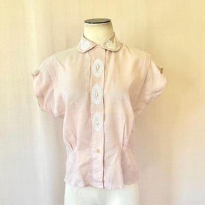 May include: A light pink short-sleeved blouse with a Peter Pan collar and decorative white embroidery. The blouse has a button-down front and is displayed on a mannequin. The fabric appears to be lightweight.