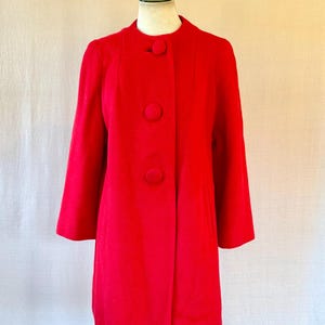 May include: A bright red coat with three large buttons. The coat has a classic design with a round collar and three-quarter length sleeves. The coat is displayed on a mannequin. The coat's length is approximately 90 cm.
