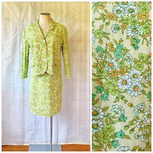 May include: A vintage two-piece suit featuring a light green floral pattern. The suit includes a collared blazer with two front pockets and a matching skirt. The fabric has a pattern of white, yellow, and green flowers.