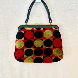 Puede incluir: Bolso de mano vintage con un patrón geométrico de octágonos rojos, negros y dorados. El bolso tiene un marco y asas de color verde oscuro. Las asas son de color verde oscuro. El bolso cuelga de un gancho blanco.