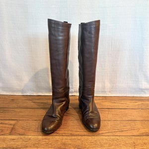 Puede incluir: Un par de botas altas de cuero marrón. Las botas son hasta la rodilla con un acabado liso y pulido. Tienen un diseño clásico con un ligero tacón y una punta redondeada. Las botas se exhiben sobre una superficie de madera.