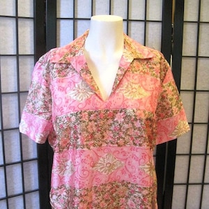May include: Pink short-sleeved blouse with a V-neck collar. The blouse features a floral pattern in shades of pink, brown, and gold. The fabric appears to be lightweight, suitable for warm weather. The blouse is displayed on a mannequin.