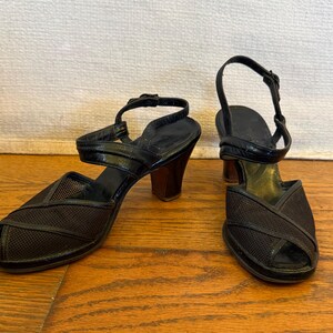 Op de afbeelding: Een paar zwarte sandalen met open teen, voorzien van een bovenwerk van mesh met kruislingse details, een hielbandje met verstelbare gesp en een stevige blokhak. Het voetbed is zwart met een goudkleurig gedeelte bij de hiel.