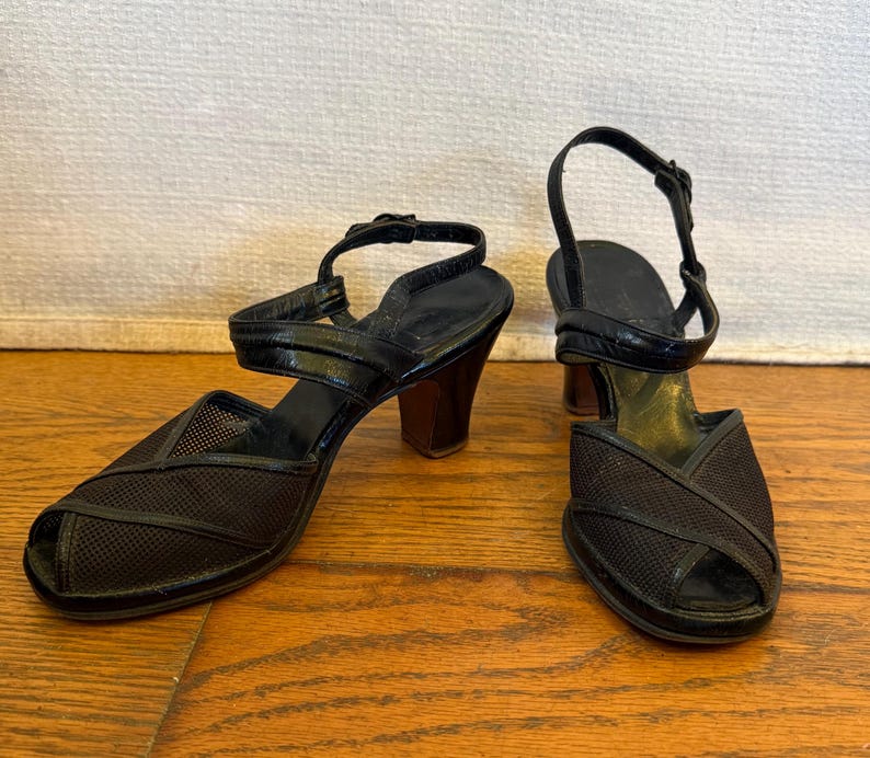 Op de afbeelding: Een paar zwarte sandalen met open teen, voorzien van een bovenwerk van mesh met kruislingse details, een hielbandje met verstelbare gesp en een stevige blokhak. Het voetbed is zwart met een goudkleurig gedeelte bij de hiel.
