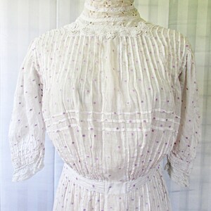 May include: A white cotton dress with a high neck and a lace collar. The dress has a dotted pattern with purple dots and is pleated throughout. The dress has 3/4 sleeves with lace trim.