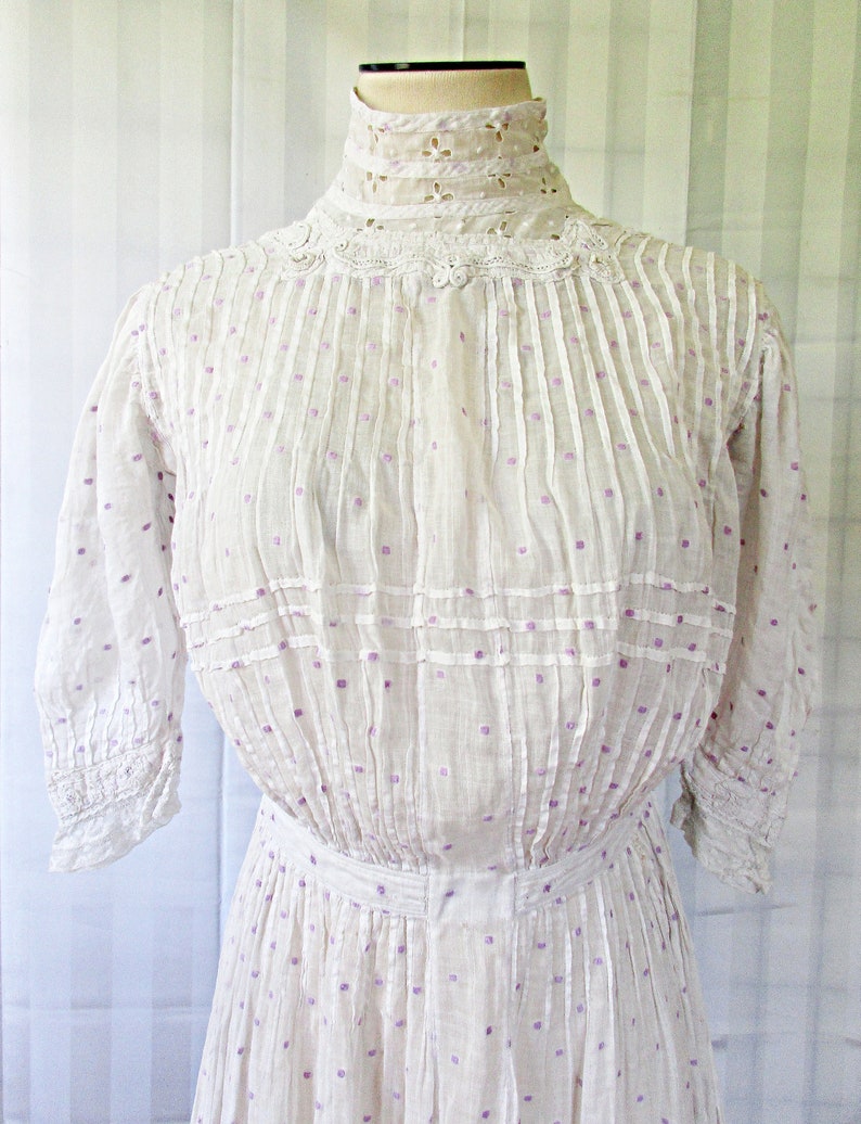 May include: A white cotton dress with a high neck and a lace collar. The dress has a dotted pattern with purple dots and is pleated throughout. The dress has 3/4 sleeves with lace trim.