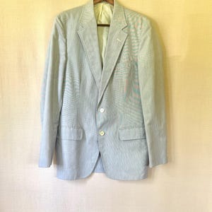 May include: A light blue blazer with a subtle striped pattern, hanging on a wooden hanger. The jacket has two front pockets, a two-button closure, and a notched lapel. The lining is a light yellow color.