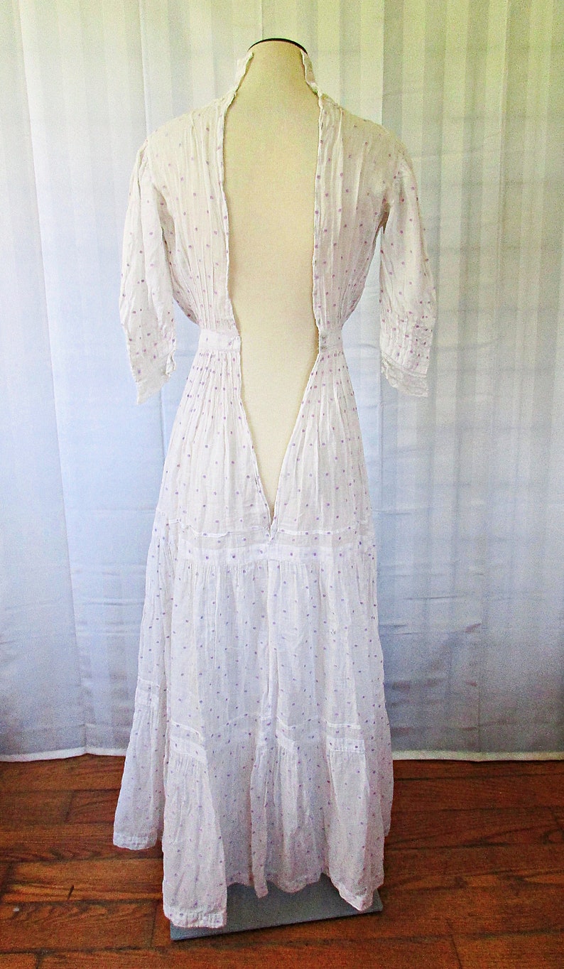 May include: A white cotton dress with a purple polka dot pattern. The dress has a deep V-neckline and a tiered skirt. The dress is on a white mannequin.