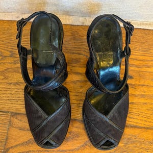 Op de afbeelding: Een paar zwarte slingback-hakken met een getextureerde mesh neus, gekruiste lederen afwerking, een verstelbare enkelband met metalen gesp en een gladde zwarte binnenzool. De schoenen hebben een open teen en een gestructureerde hak.
