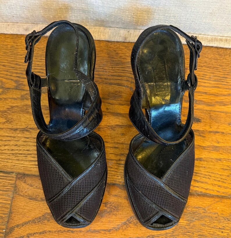 Op de afbeelding: Een paar zwarte slingback-hakken met een getextureerde mesh neus, gekruiste lederen afwerking, een verstelbare enkelband met metalen gesp en een gladde zwarte binnenzool. De schoenen hebben een open teen en een gestructureerde hak.