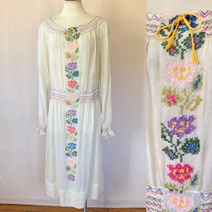 Può includere: Un abito vintage bianco con maniche lunghe e un ricamo floreale. Il ricamo presenta fiori e foglie colorati in un motivo verticale sul davanti dell'abito.