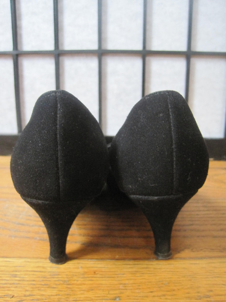 Op de afbeelding: Een paar zwarte su&egrave;de pumps met een lage, afgeronde hak. De schoenen hebben een klassiek ontwerp, met een spitse neus en een gladde afwerking. De hakken zijn ongeveer 5 cm hoog.