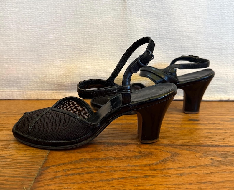Op de afbeelding: Een paar zwarte schoenen met open teen, voorzien van een bovenwerk van mesh-stof, een afgeronde neus en een dun enkelbandje met gespsluiting. De schoenen hebben een effen zwarte hak en een glad voetbed, met duidelijk zichtbare stiksels.