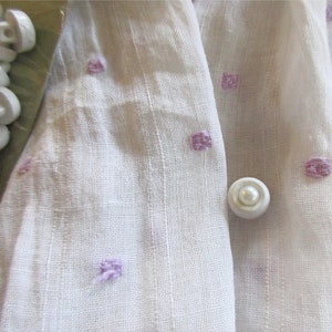 May include: White fabric with a subtle texture and a pattern of small, light purple squares. A single white pearl button is centered in the image.
