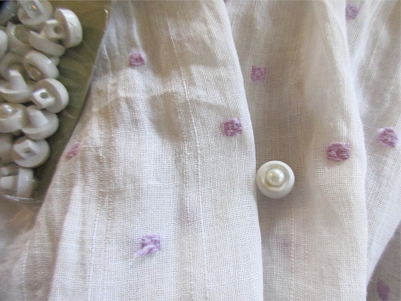 May include: White fabric with a subtle texture and a pattern of small, light purple squares. A single white pearl button is centered in the image.