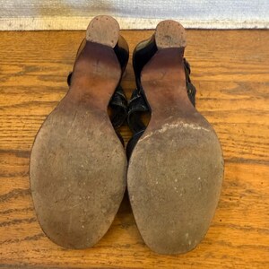 Op de afbeelding: Een paar zwarte sandalen met houten hakken en zolen, gezien vanaf de onderkant. De zolen zijn versleten en getextureerd, met een afgeronde neus en een stevige blokhak. De bovenste bandjes zijn gedeeltelijk zichtbaar in zwart materiaal.