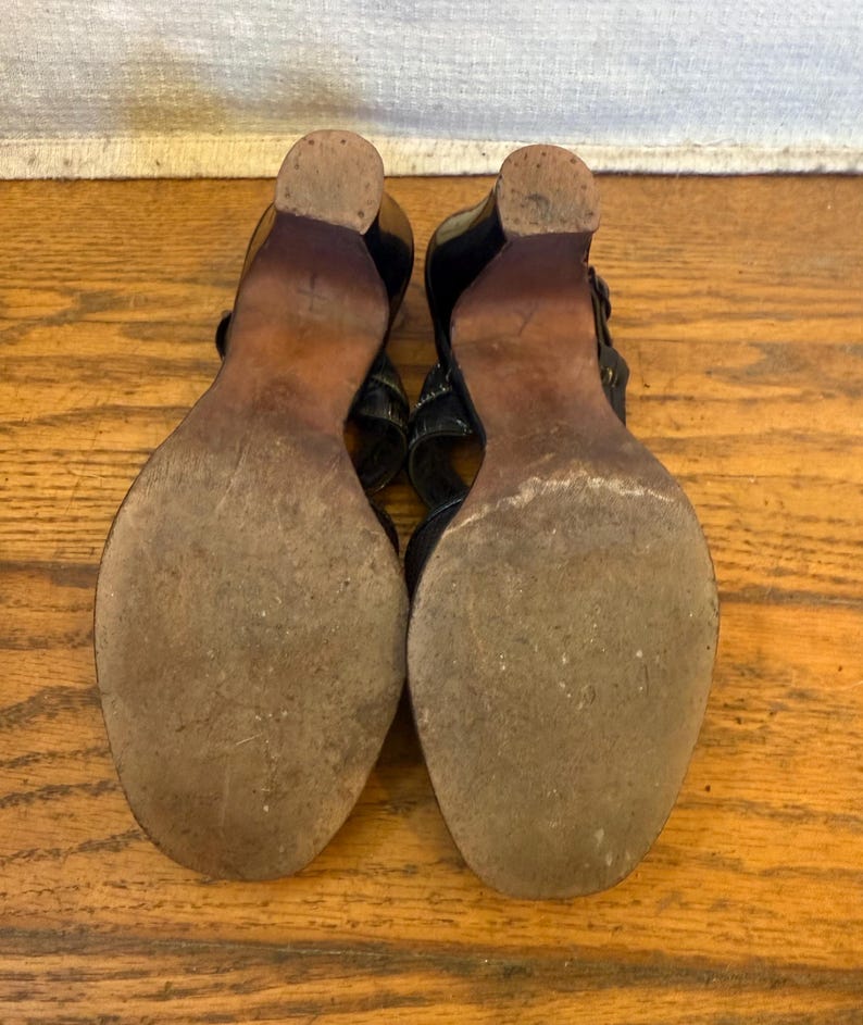 Op de afbeelding: Een paar zwarte sandalen met houten hakken en zolen, gezien vanaf de onderkant. De zolen zijn versleten en getextureerd, met een afgeronde neus en een stevige blokhak. De bovenste bandjes zijn gedeeltelijk zichtbaar in zwart materiaal.