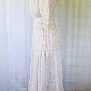 May include: A white cotton dress with a high neck and a gathered bodice. The dress has a dotted pattern with pink and purple dots. The dress has a tiered skirt and is fitted at the waist.