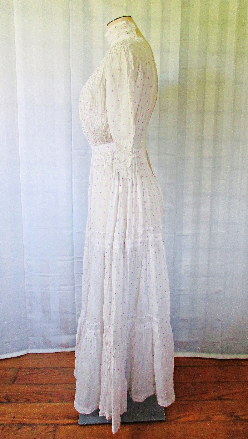 May include: A white cotton dress with a high neck and a gathered bodice. The dress has a dotted pattern with pink and purple dots. The dress has a tiered skirt and is fitted at the waist.