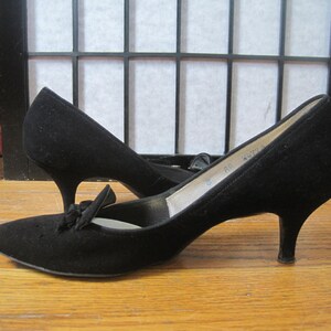 Op de afbeelding: Een paar zwarte su&egrave;de pumps met een strikdetail op de wreef. De schoenen hebben een spitse neus en een slanke, lage hak. De hakken meten ongeveer 5 cm.