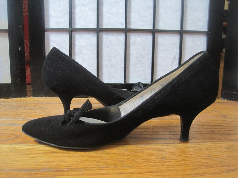 Op de afbeelding: Een paar zwarte su&egrave;de pumps met een strikdetail op de wreef. De schoenen hebben een spitse neus en een slanke, lage hak. De hakken meten ongeveer 5 cm.
