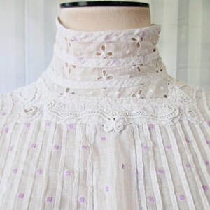 May include: White cotton blouse with a high, lace collar and a delicate pattern of purple dots. The blouse has a gathered neckline and a button closure at the front.