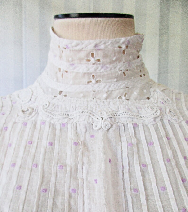 May include: White cotton blouse with a high, lace collar and a delicate pattern of purple dots. The blouse has a gathered neckline and a button closure at the front.