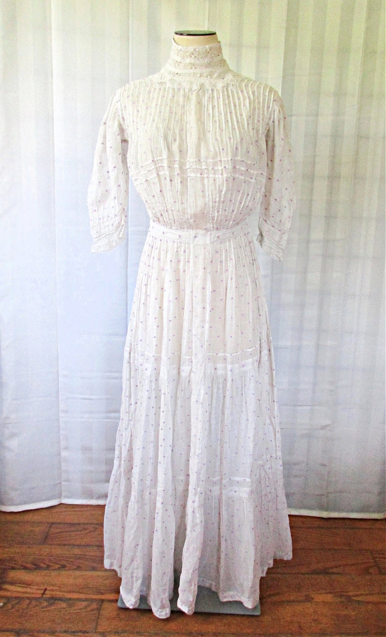 May include: A white cotton dress with a high neck and a gathered skirt. The dress has a small purple polka dot pattern and is decorated with lace trim.