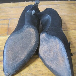 Op de afbeelding: Een paar zwarte su&egrave;de pumps. De schoenen hebben spitse tenen en stilettohakken. De zolen vertonen slijtage. De hakken zijn ongeveer 7.6 cm hoog.