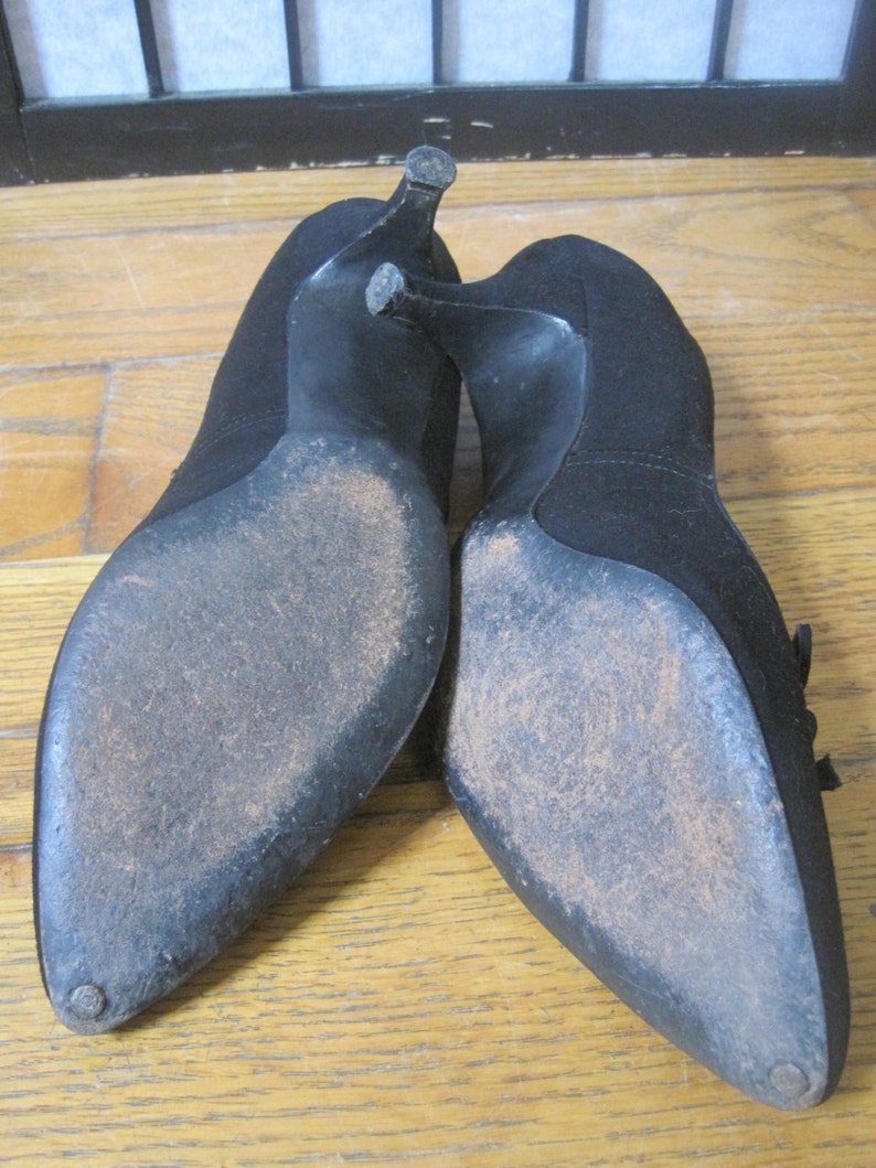 Op de afbeelding: Een paar zwarte su&egrave;de pumps. De schoenen hebben spitse tenen en stilettohakken. De zolen vertonen slijtage. De hakken zijn ongeveer 7.6 cm hoog.