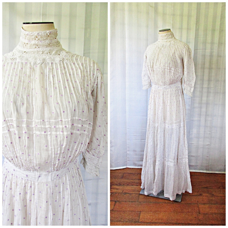 May include: A white cotton dress with a high neck and long sleeves. The dress has a white lace trim around the neck and a white lace overlay on the bodice. The dress is covered in small purple polka dots and has a tiered skirt.