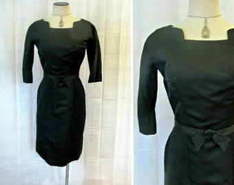 Può includere: Un abito vintage nero con scollo quadrato, maniche a tre quarti e silhouette aderente. L'abito ha una cintura con un fiocco in vita.