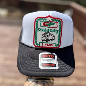 May include: A black and white trucker hat with a white front panel and a black mesh back. The front features a patch with the text "Shield of Safety" and a truck graphic. The hat also has a "OTTO" tag.