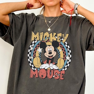 Puede incluir: Una camiseta gris carb&oacute;n de manga corta con un gr&aacute;fico de Mickey Mouse centrado en un marco circular. El dise&ntilde;o incluye el texto MICKEY MOUSE en letras negritas, un borde a cuadros y detalles de estrellas en tonos amarillo, rojo y azul.